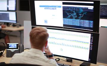 Male Viasat employee sitting at a desk in front of two stacked monitors in a cybersecurity operations center