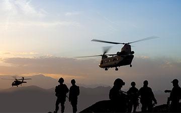 Silhouettes of six army men in the field at dusk with two helicopters flying away
