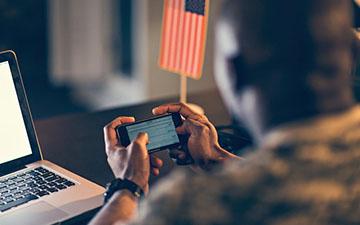 Close-up of a man in the military holding a smartphone horizontally in front of a laptop and american flag on a stand