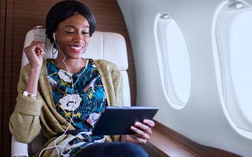 Professional woman with black hair sitting on a private plane, listening to content on a tablet with earbuds